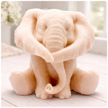 Load image into Gallery viewer, A crisp product photograph of a detailed, grey artisan bar of soap intricately molded into the shape of a sitting baby elephant. The elephant is depicted holding its trunk with its front legs crossed in a "hear no evil" or hugging pose, showcasing realistic skin textures, expressive eyes, and large ears. The product is perfectly centered against a seamless, solid, plain white background.
