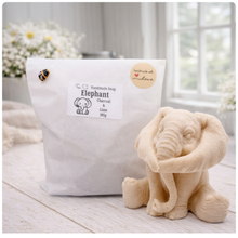 Load image into Gallery viewer, A crisp product photograph of a detailed, grey artisan bar of soap intricately molded into the shape of a sitting baby elephant. The elephant is depicted holding its trunk with its front legs crossed in a "hear no evil" or hugging pose, showcasing realistic skin textures, expressive eyes, and large ears. The product is perfectly centered against a seamless, solid, plain white background.
