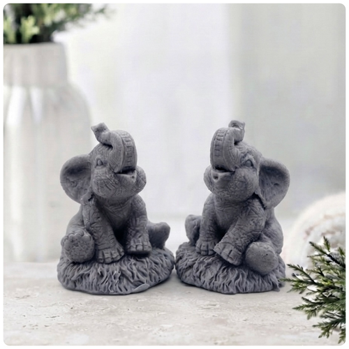 A charming gift set of two handmade vegan soaps, one sculpted as a sitting baby elephant with a trunk raised in a greeting and the other as a smaller standing calf, displayed on a light wooden surface in a bright farmhouse setting.