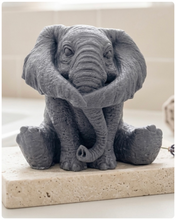Load image into Gallery viewer, A highly detailed handmade vegan soap in a charcoal grey tone, sculpted in the shape of a sitting elephant with realistic wrinkled skin textures and large ears, displayed on a light wooden surface with white flowers in the background.
