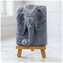 Load image into Gallery viewer, A rectangular handmade vegan soap in a speckled slate-grey finish, featuring a highly detailed relief of an elephant's head adorned with ornate, traditional patterns and a decorative floral background, displayed on a miniature wooden easel.
