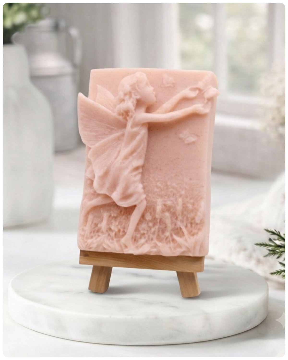 A rectangular handmade vegan soap featuring a detailed relief of a winged fairy reaching towards a butterfly in a meadow of tall grass and flowers, displayed on a miniature wooden easel
