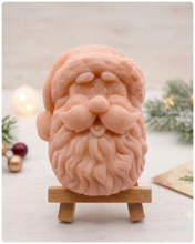 Load image into Gallery viewer, handmade vegan soap sculpted in the shape of Father Christmas's head, featuring a detailed curly beard, a jolly moustache, and a classic Santa hat, displayed on a miniature wooden easel in a bright farmhouse setting.
