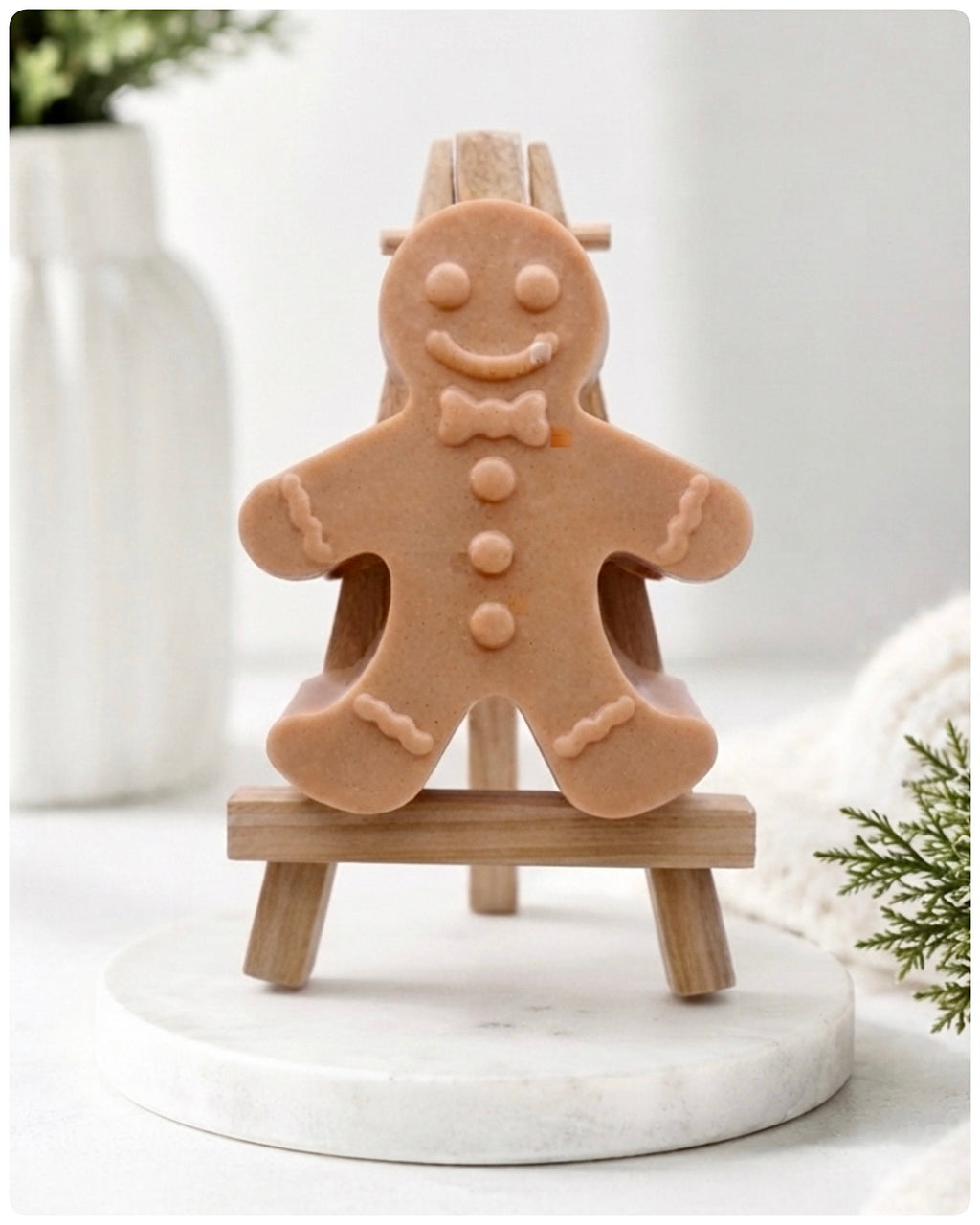 A handmade vegan featuring a classic gingerbread man with a smiling face, a bow tie, and decorative icing details on his arms and legs, displayed on a miniature wooden easel.