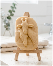 Load image into Gallery viewer, A rectangular handmade vegan soap in a natural tone, featuring a detailed relief of a mother and baby giraffe with their necks intertwined, displayed on a miniature wooden easel against a white wood-panelled background.
