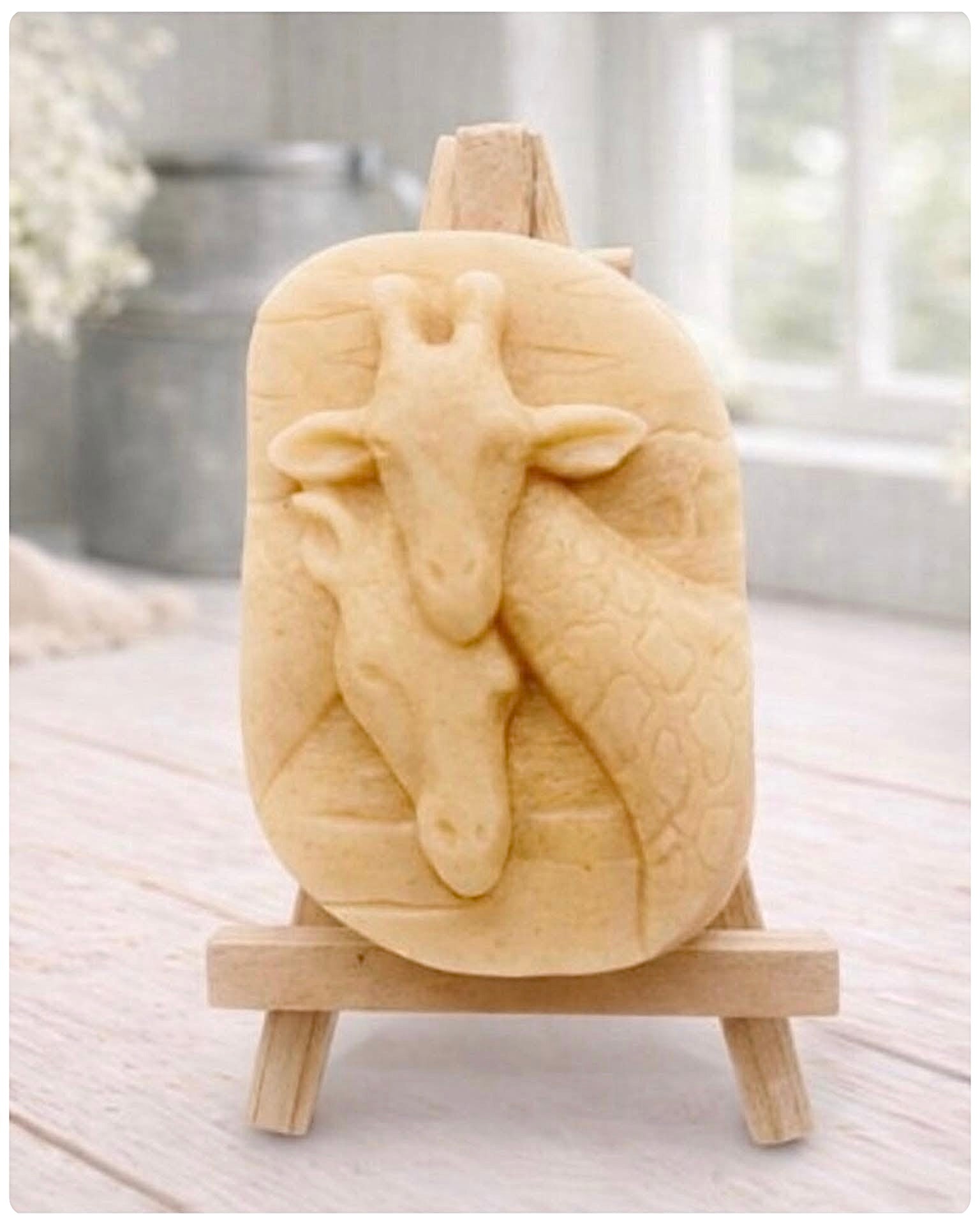 A rectangular handmade vegan soap in a natural tone, featuring a detailed relief of a mother and baby giraffe with their necks intertwined, displayed on a miniature wooden easel against a white wood-panelled background.