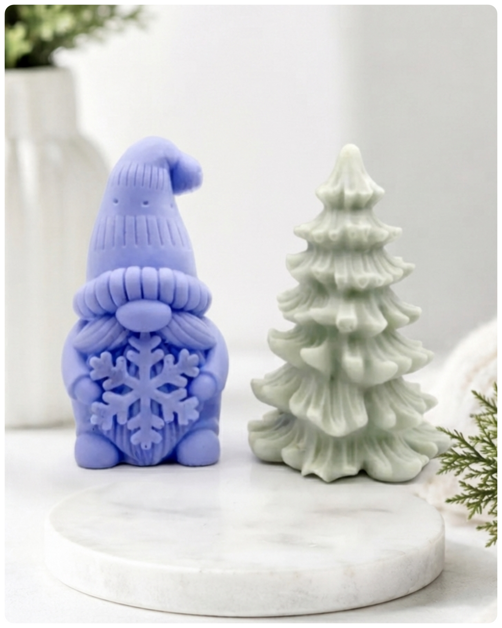 A pair of handmade vegan soaps featuring a Christmas gonk (gnome) with a textured hat holding a large snowflake, standing next to a pale mint-green 3D Christmas tree with layered, snow-dusted branches, displayed in a bright farmhouse setting.