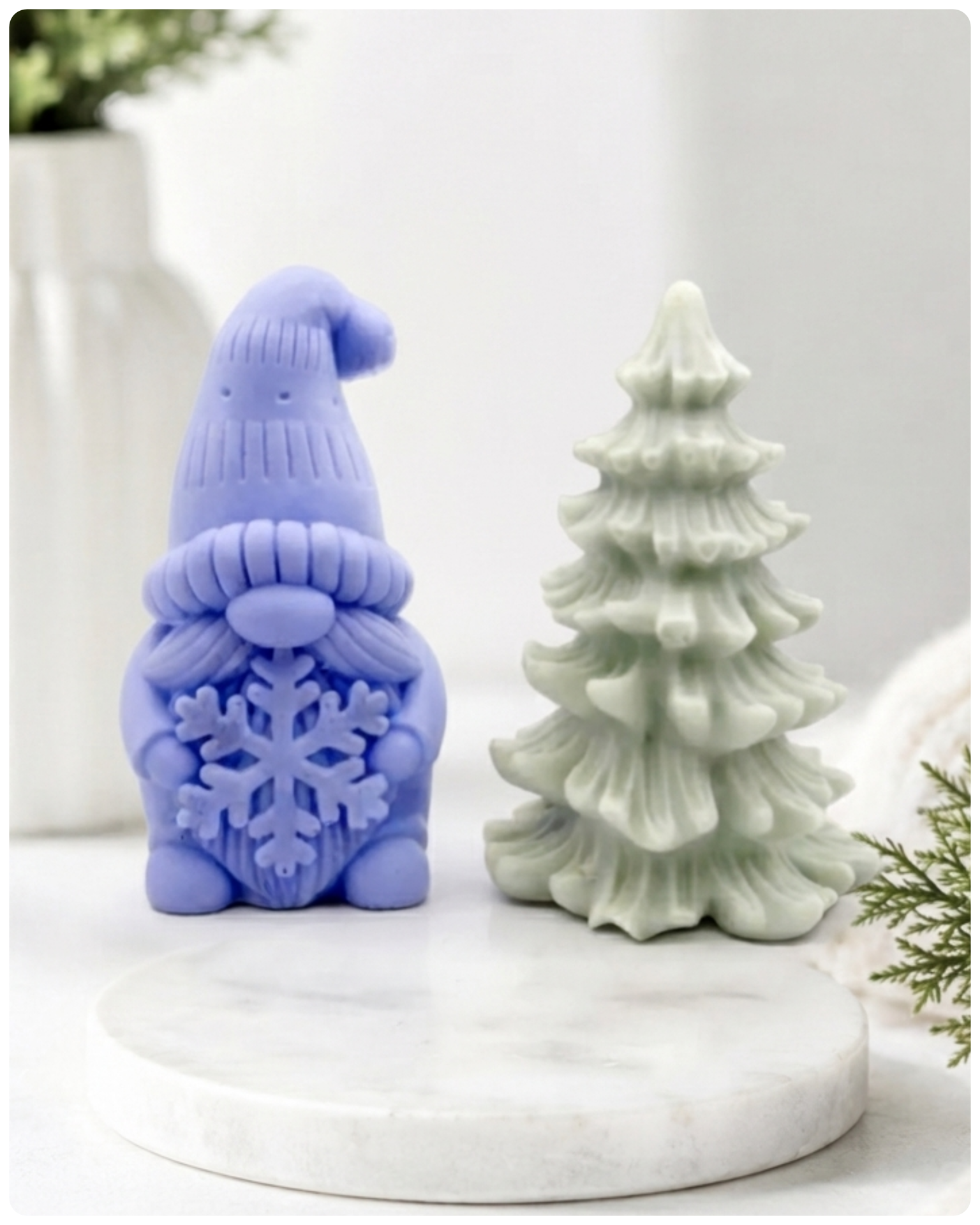 A pair of handmade vegan soaps featuring a Christmas gonk (gnome) with a textured hat holding a large snowflake, standing next to a pale mint-green 3D Christmas tree with layered, snow-dusted branches, displayed in a bright farmhouse setting.