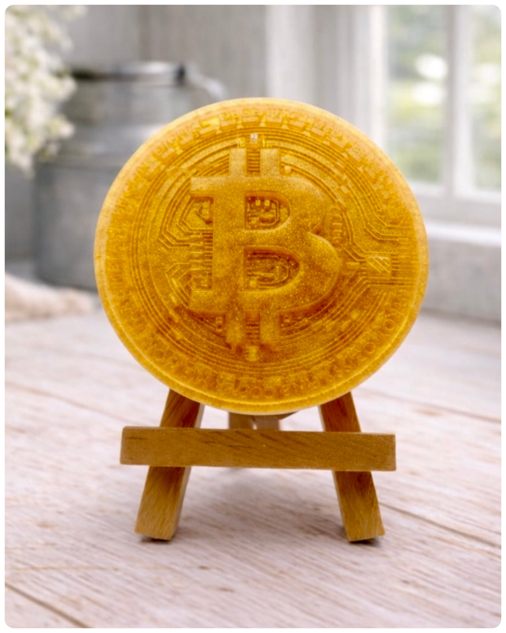handmade vegan soap in the shape of a Bitcoin coin with a shimmering glitter finish, featuring a detailed relief of the "B" symbol and circuit board patterns, displayed on a small wooden easel in a bright farmhouse decor setting.