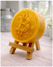 Load image into Gallery viewer, handmade vegan soap in the shape of a Bitcoin coin with a shimmering glitter finish, featuring a detailed relief of the "B" symbol and circuit board patterns, displayed on a small wooden easel in a bright farmhouse decor setting.
