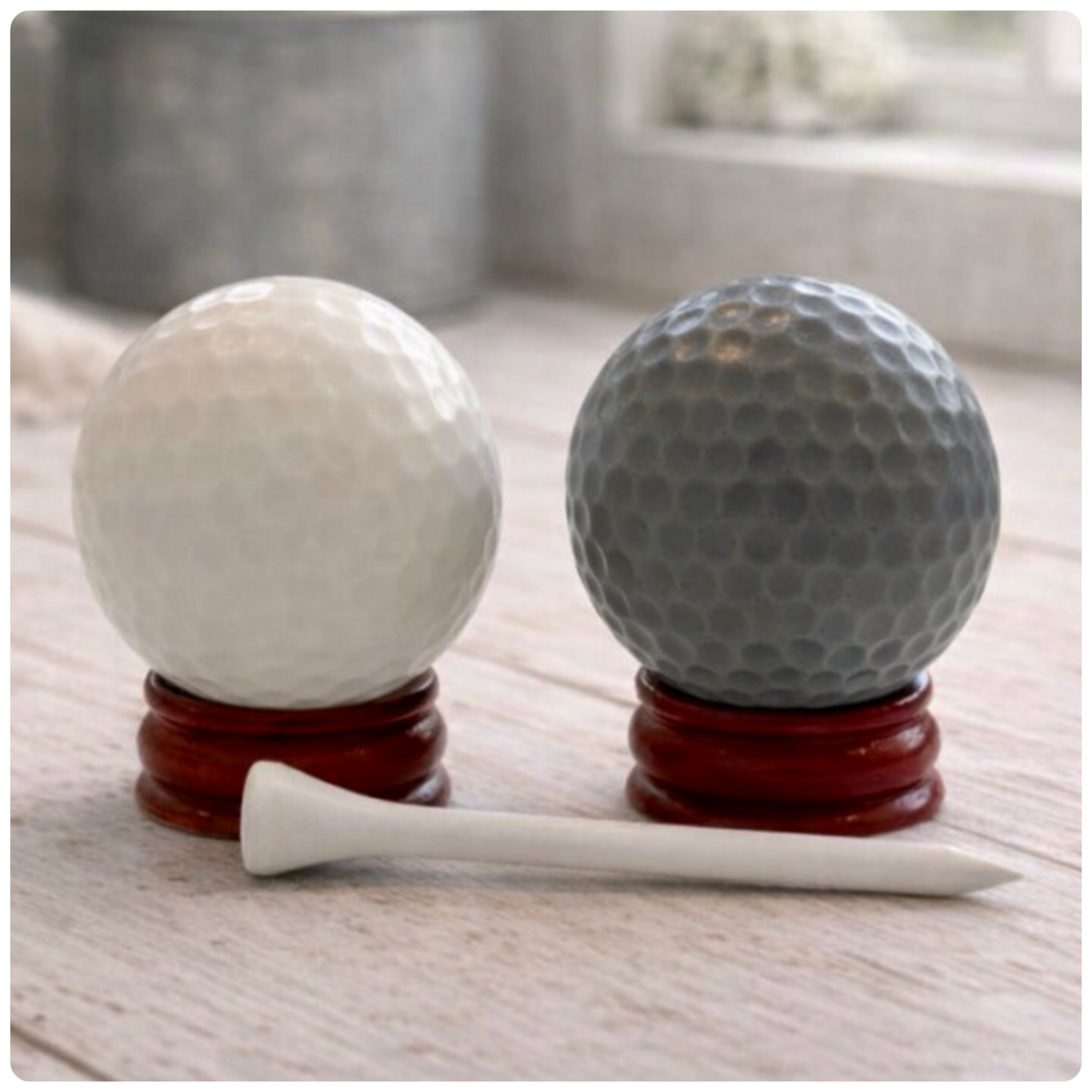 A handmade vegan golf ball soap gift set featuring one white and one grey spherical soap with realistic dimpled textures, displayed on small wooden stands next to a white golf tee and a kraft gift box tied with a red ribbon