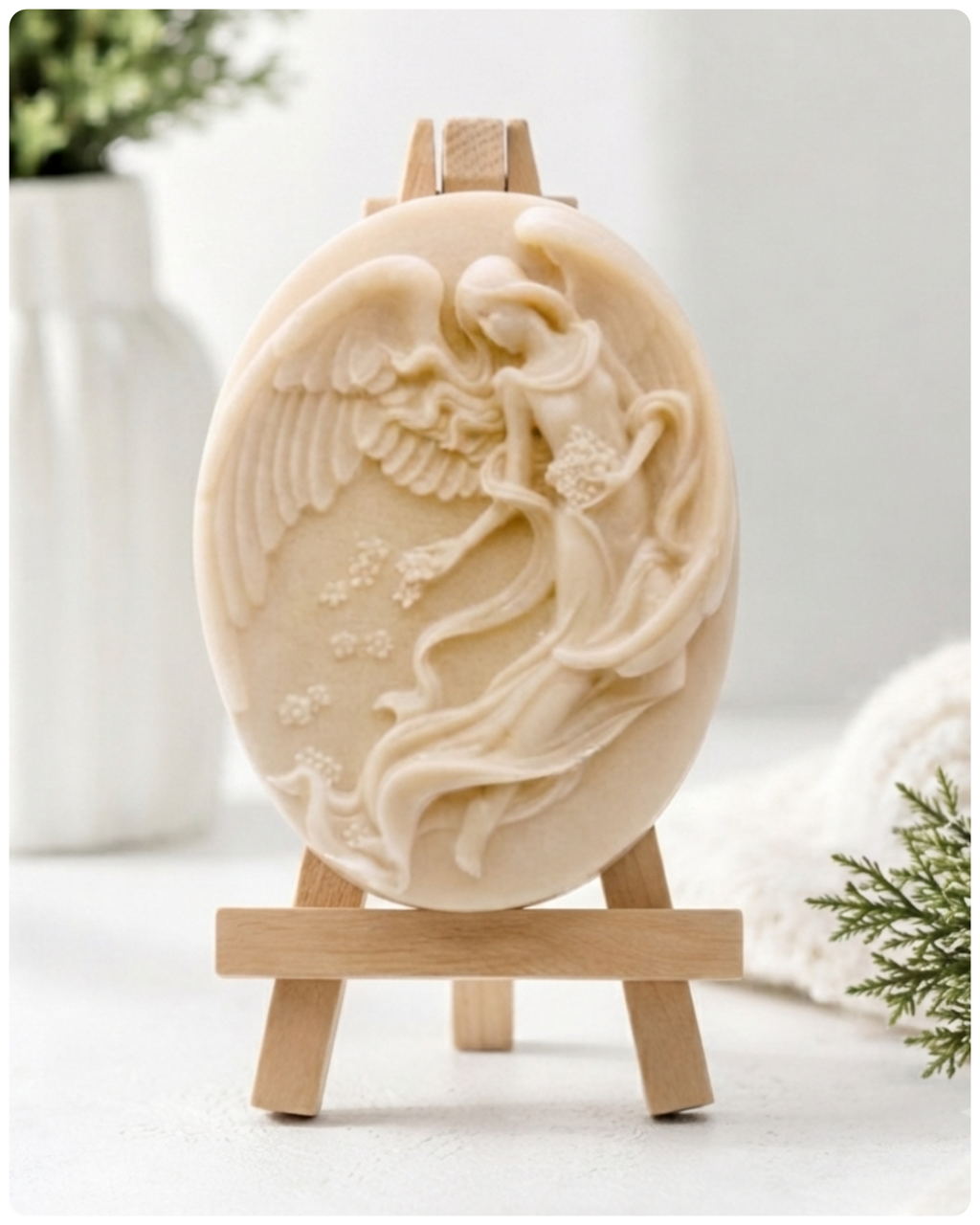 An oval-shaped handmade vegan soap, featuring an intricate relief carving of a guardian angel with large wings holding flowers, displayed on a small wooden easel.