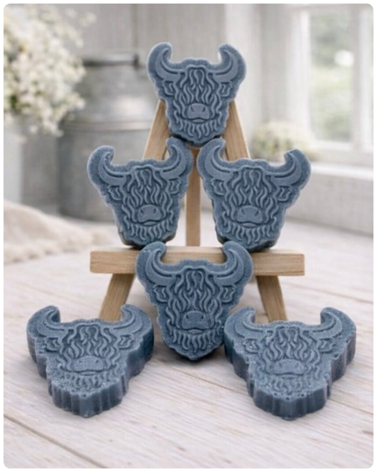 A collection of six handmade vegan soaps each intricately sculpted into the head of a shaggy Highland cow with long horns and a thick, textured coat, arranged in a pyramid on and around a small wooden easel.