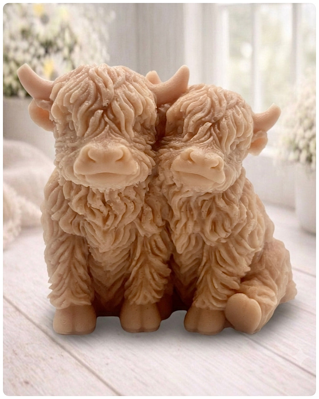 Handmade soap featuring two Highland cows with textured, shaggy fur sitting side by side on a light wooden surface with a softly indoor background.