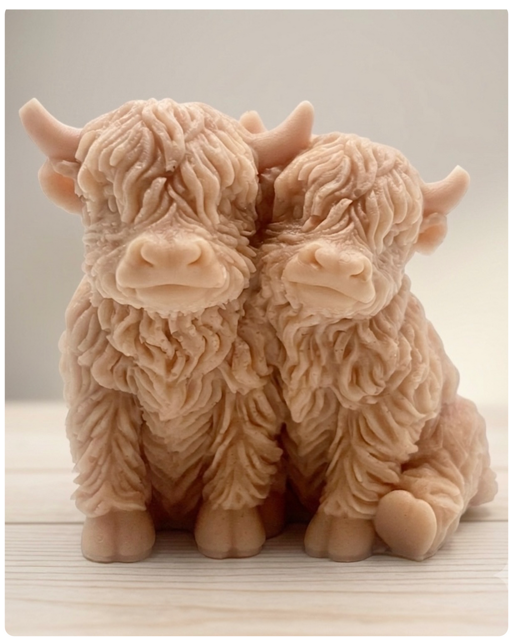 Handmade soap featuring two Highland cows with textured, shaggy fur sitting side by side on a light wooden surface with a softly indoor background.