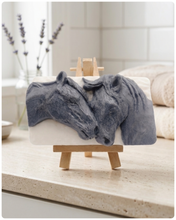 Load image into Gallery viewer, A rectangular handmade vegan soap featuring a detailed relief of two horse heads leaning affectionately towards each other, finished in a slate-grey tone against a white soap base and displayed on a  wooden easel.
