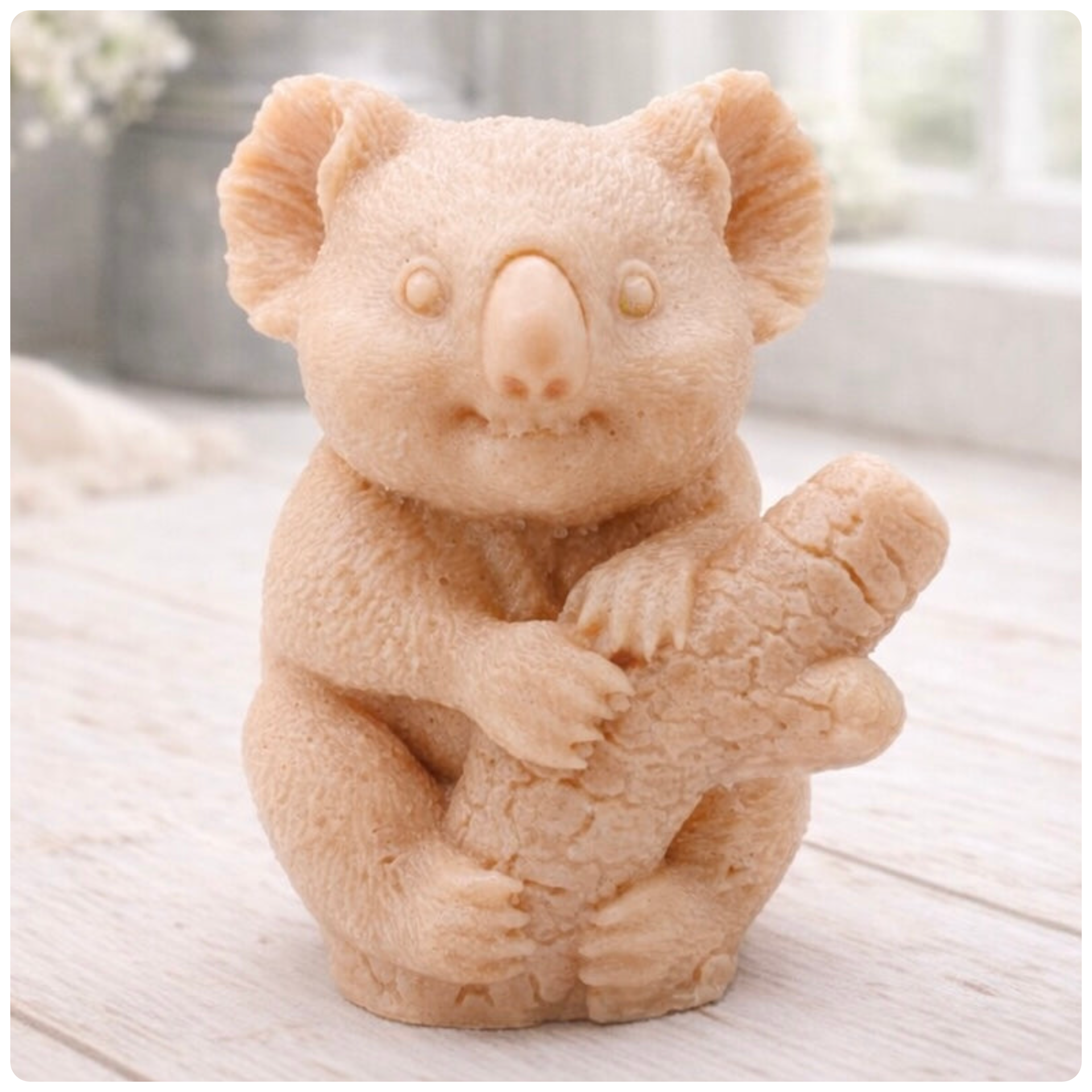 A highly detailed handmade vegan soap in a sculpted in the shape of a realistic koala bear sitting upright and clutching a textured eucalyptus branch, displayed on a light wooden surface in a bright farmhouse background 