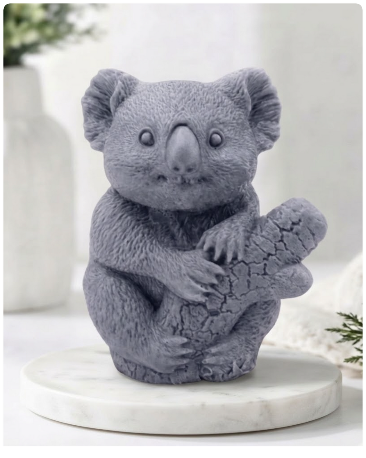 A front-facing photograph of a grey, charcoal-coloured koala-shaped soap bar. The highly detailed soap is sculpted to show textured fur and the koala clinging to a textured log. It sits on a round, white marble disc. In the blurry background, there is a white ceramic vase with green sprigs and a rolled white towel.