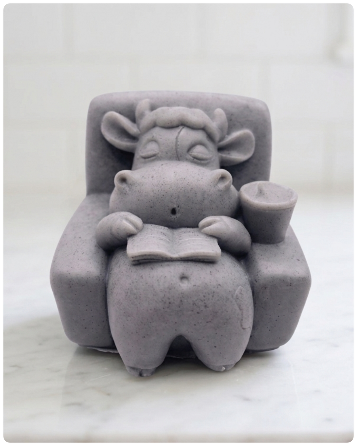 A handmade vegan soap sculpted to show a whimsical "lazy cow" character relaxing in an armchair, reading a book and holding a drink,