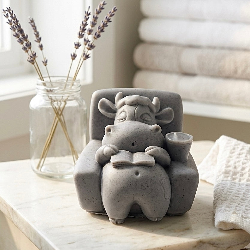 A handmade vegan soap sculpted to show a whimsical "lazy cow" character relaxing in an armchair, reading a book and holding a drink,
