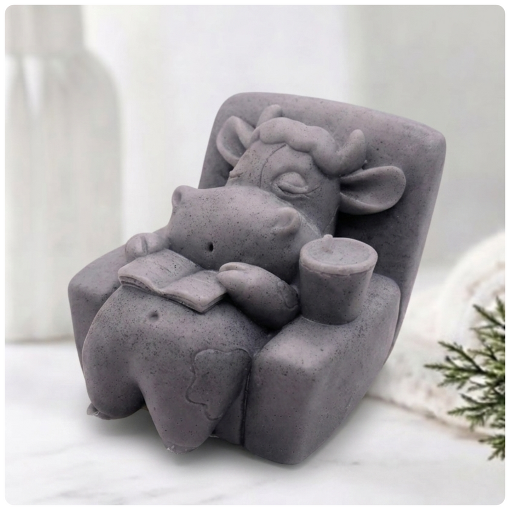 Handmade grey Lazy Cow soap sitting in an armchair reading a book with a candle, eyes closed in a relaxed pose. Unique decorative animal soap by Soap Simple Creations, Blackpool UK.