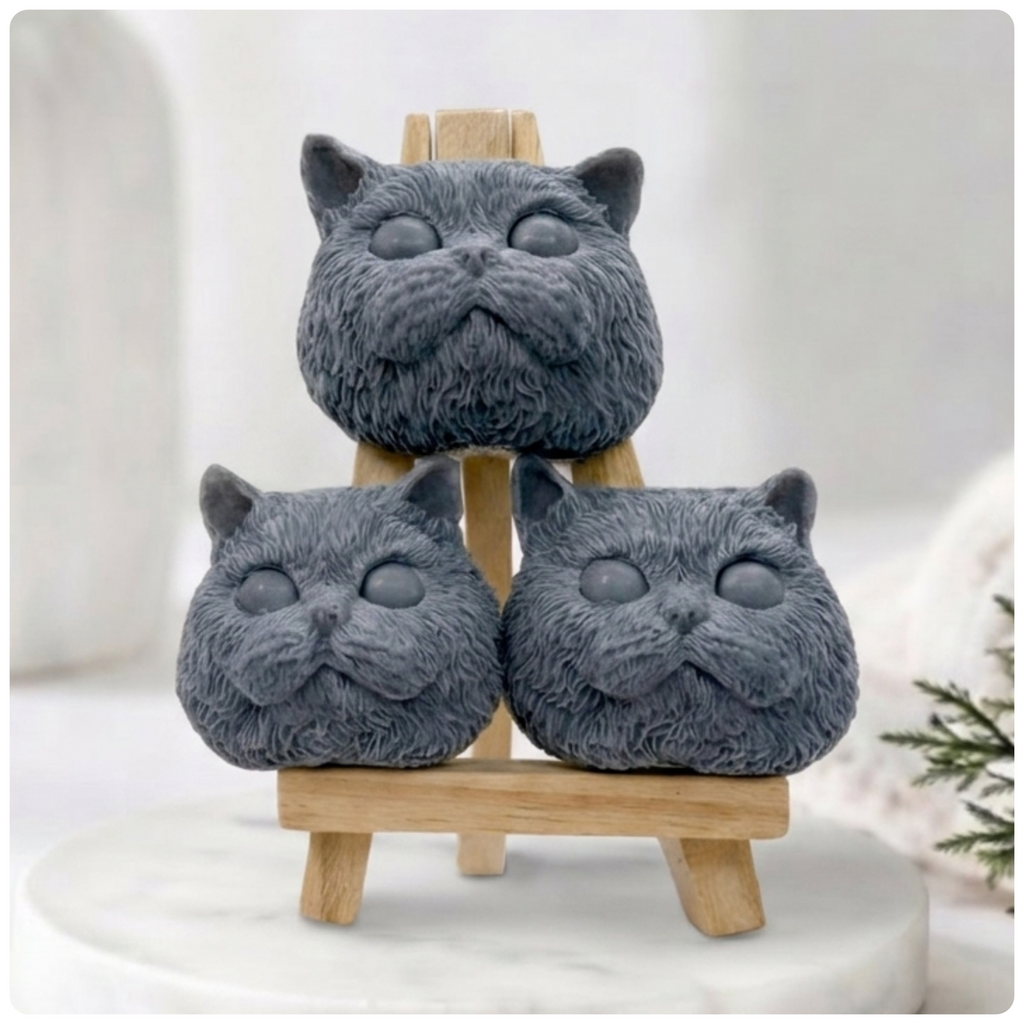 Three dark grey handmade cat face soaps featuring round eyes, chubby cheeks, and detailed fur texture stacked on a small wooden display easel on a marble coaster. Gift boxed 
