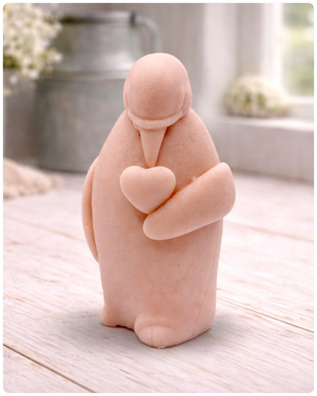 A handmade vegan soap sculpted as a standing penguin tenderly cradling a heart against its chest, displayed on a light wooden surface in a bright farmhouse setting.