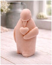 Load image into Gallery viewer, A handmade vegan soap sculpted as a standing penguin tenderly cradling a heart against its chest, displayed on a light wooden surface in a bright farmhouse setting.
