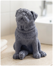 Load image into Gallery viewer, A detailed handmade vegan soap sculpted in the shape of a sitting pug dog with characteristic facial wrinkles and a tilted head, in a natural tone, displayed on a light wooden surface.
