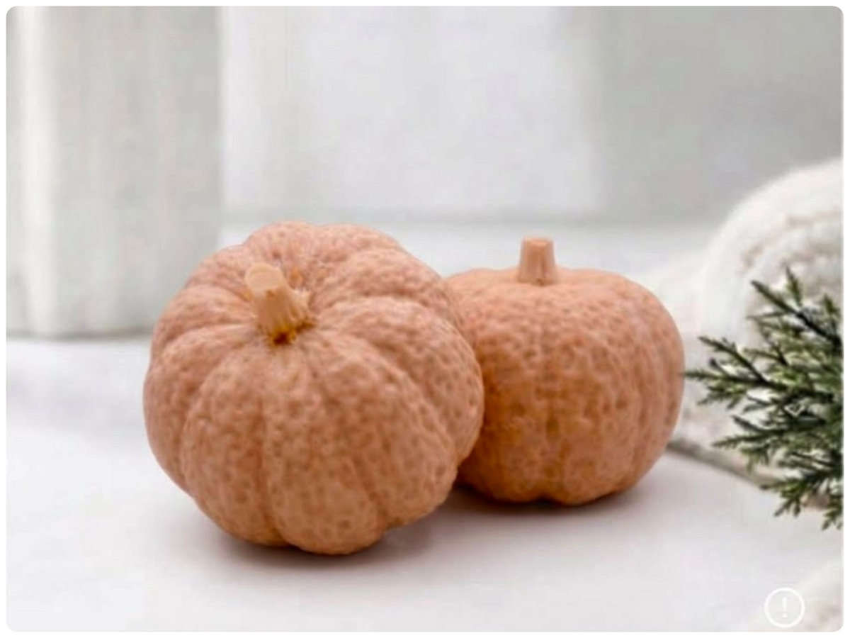 Two handmade pumpkin-shaped soaps in a warm beige peach shade, sitting on a clean surface with a white vase and greenery in the background.