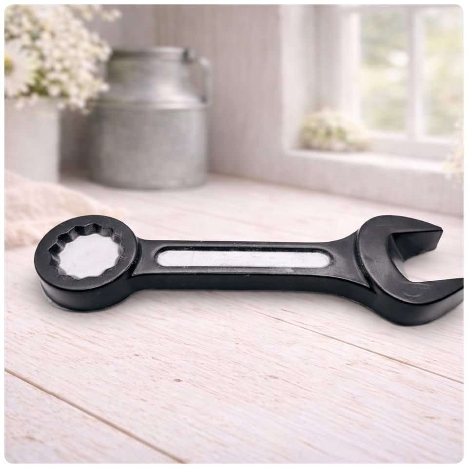 A 3D handmade vegan soap sculpted in the shape of a realistic spanner or wrench tool, finished in a sleek charcoal black with silver-painted detailing on the handle and head, lying on a light wood surface.