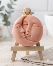 Load image into Gallery viewer, handmade vegan soap gift featuring a circular relief sculpture of a crescent moon and sun with peaceful intertwined faces, displayed on a small wooden easel in a bright farmhouse decor setting.
