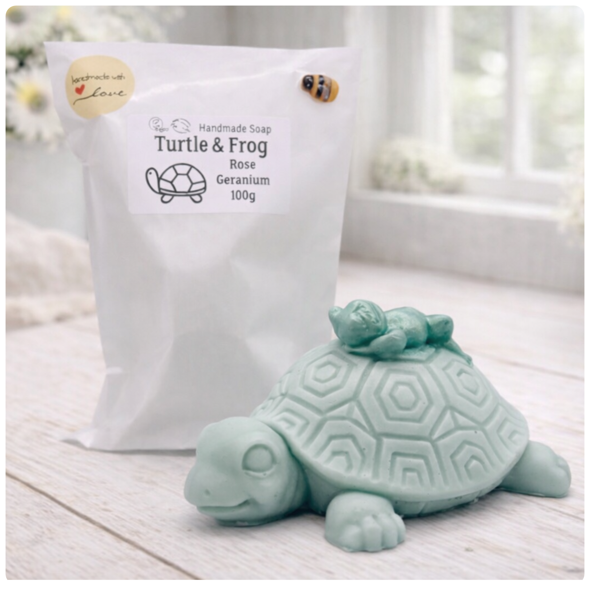 A handmade vegan soap in a soft mint green tone, sculpted into a large friendly turtle with a geometric shell pattern and a small, cute frog sleeping peacefully on its back.