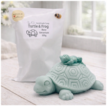 Load image into Gallery viewer, A handmade vegan soap in a soft mint green tone, sculpted into a large friendly turtle with a geometric shell pattern and a small, cute frog sleeping peacefully on its back.
