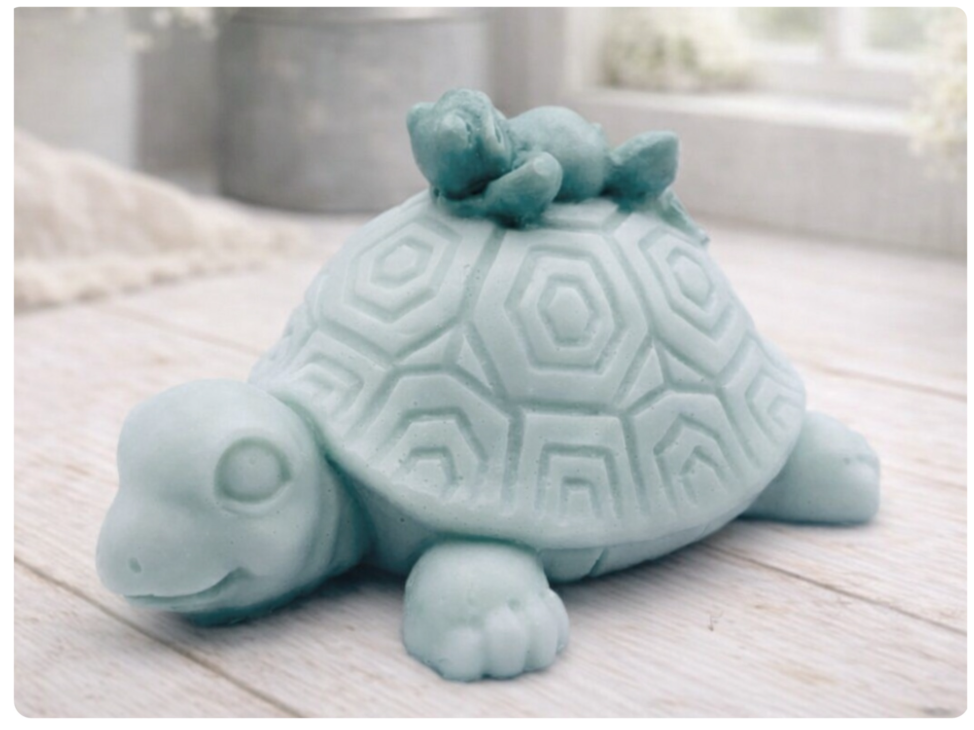 A handmade vegan soap in a soft mint green tone, sculpted into a large friendly turtle with a geometric shell pattern and a small, cute frog sleeping peacefully on its back.
