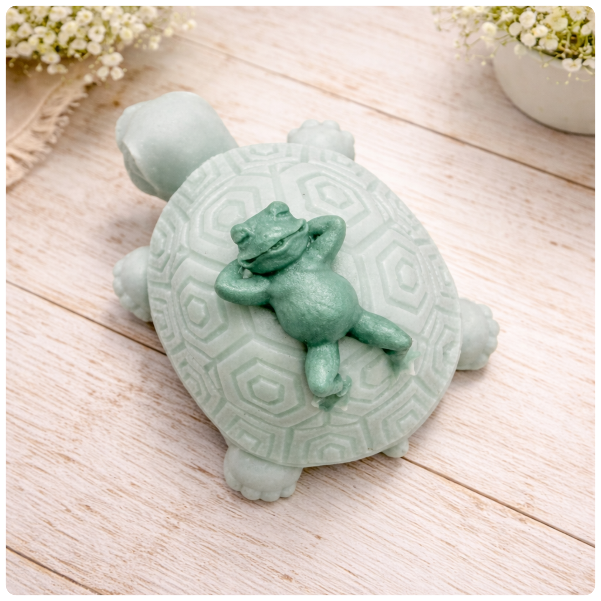 A handmade vegan soap in a soft mint green tone, sculpted into a large friendly turtle with a geometric shell pattern and a small, cute frog sleeping peacefully on its back.