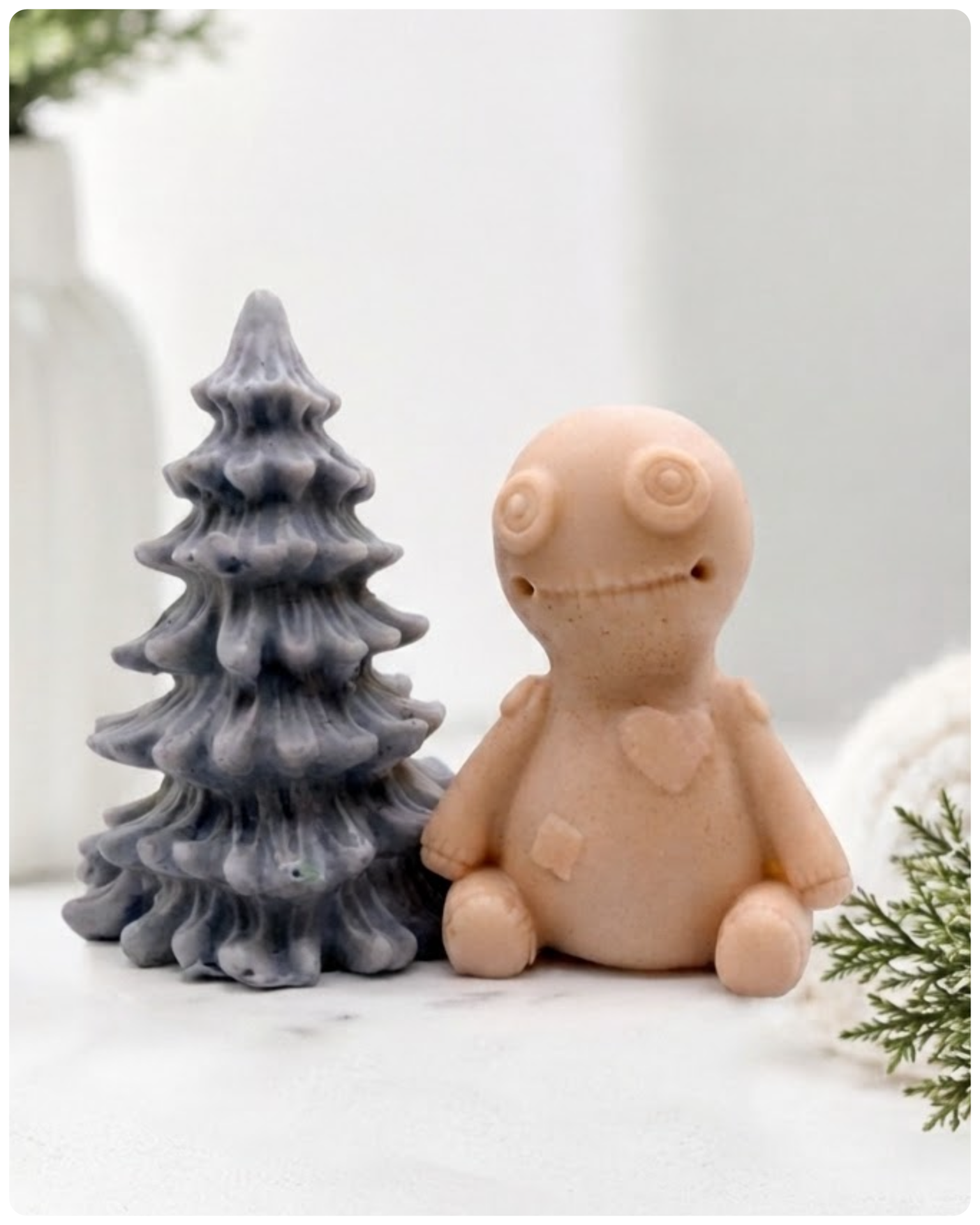 A handmade vegan soap gift set featuring a cream-coloured voodoo doll soap with stitched details and a grey tiered Christmas tree soap, presented in a black gift box tied with a red raffia ribbon and a small ladybird embellishment.