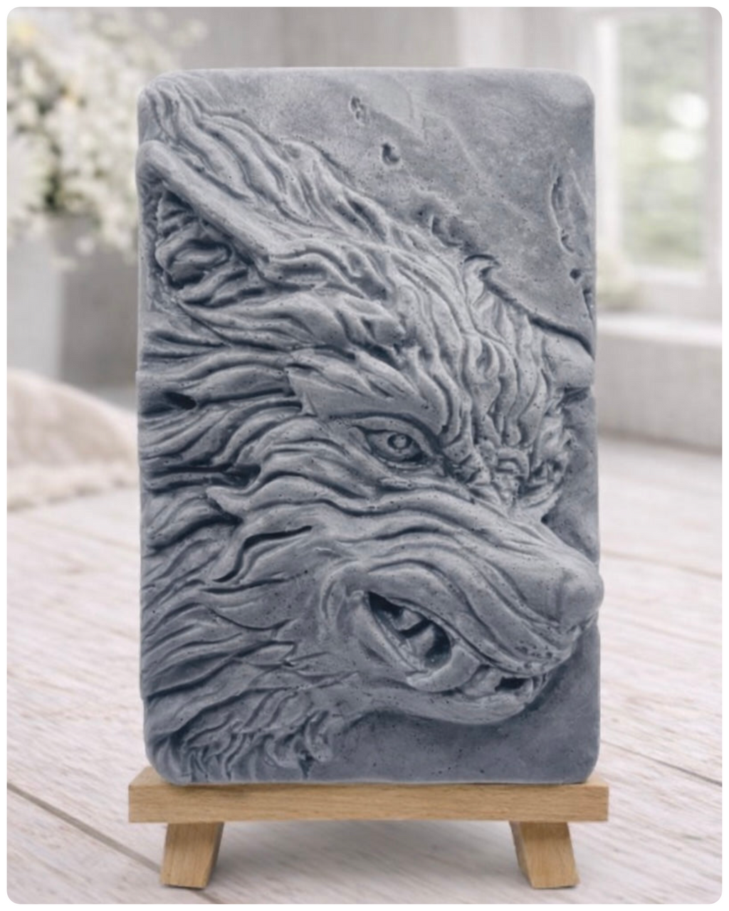 A rectangular handmade vegan soap featuring a highly detailed, 3D relief carving of a wolf's head in profile with a thick, textured mane, displayed on a miniature wooden easel.