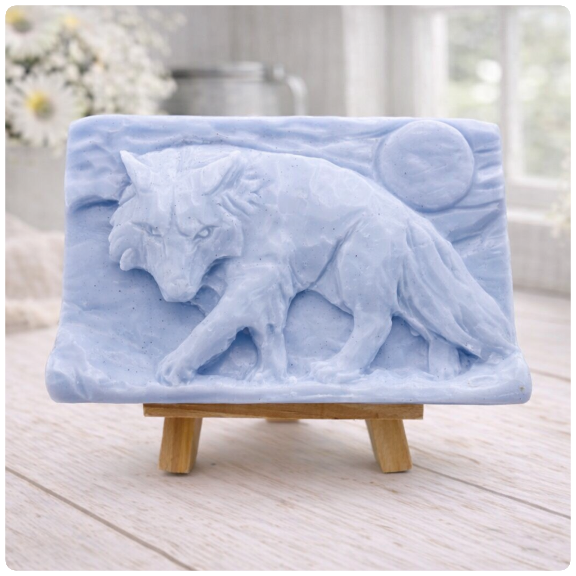 A rectangular handmade vegan soap, featuring a detailed relief carving of a wolf in mid-stride across a rugged landscape with a large full moon in the background, displayed on a miniature wooden easel.