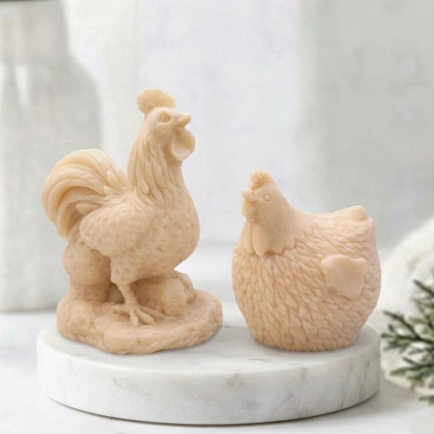Side profile of two meticulously detailed, soft yellow, handmade vegan soap bars - one cockerel and one hen - sitting on a white marble coaster against a bokeh background of a window and blurry greenery.