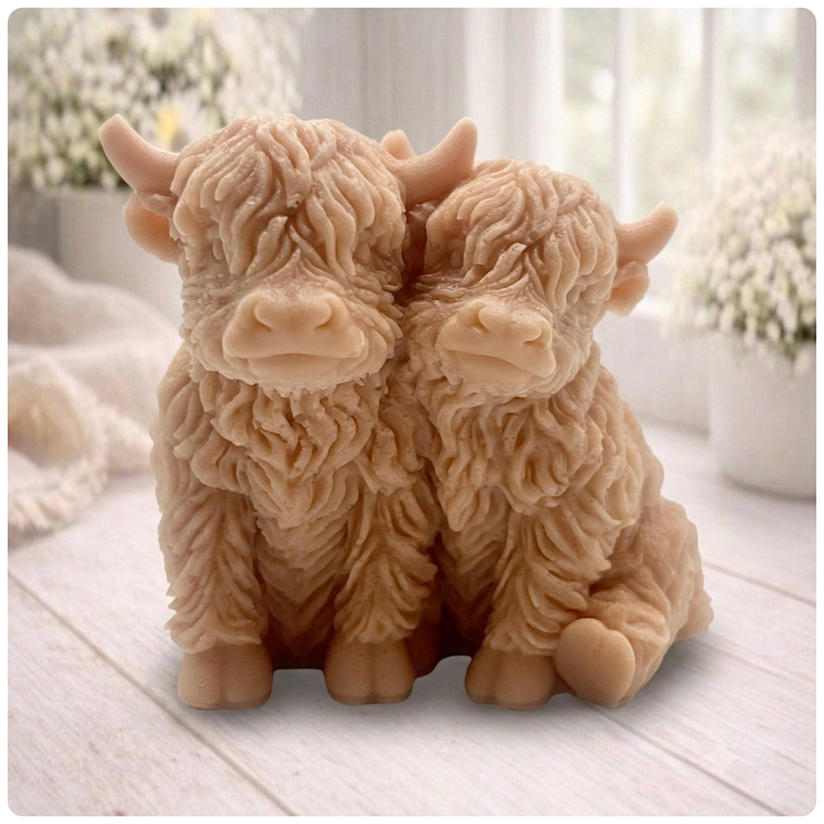 Handmade soap featuring two Highland cows with textured, shaggy fur sitting side by side on a light wooden surface with a softly indoor background.