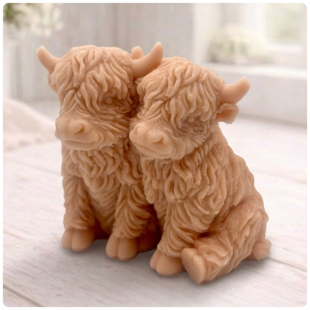 Handmade soap featuring two Highland cows with textured, shaggy fur sitting side by side on a light wooden surface with a softly indoor background.