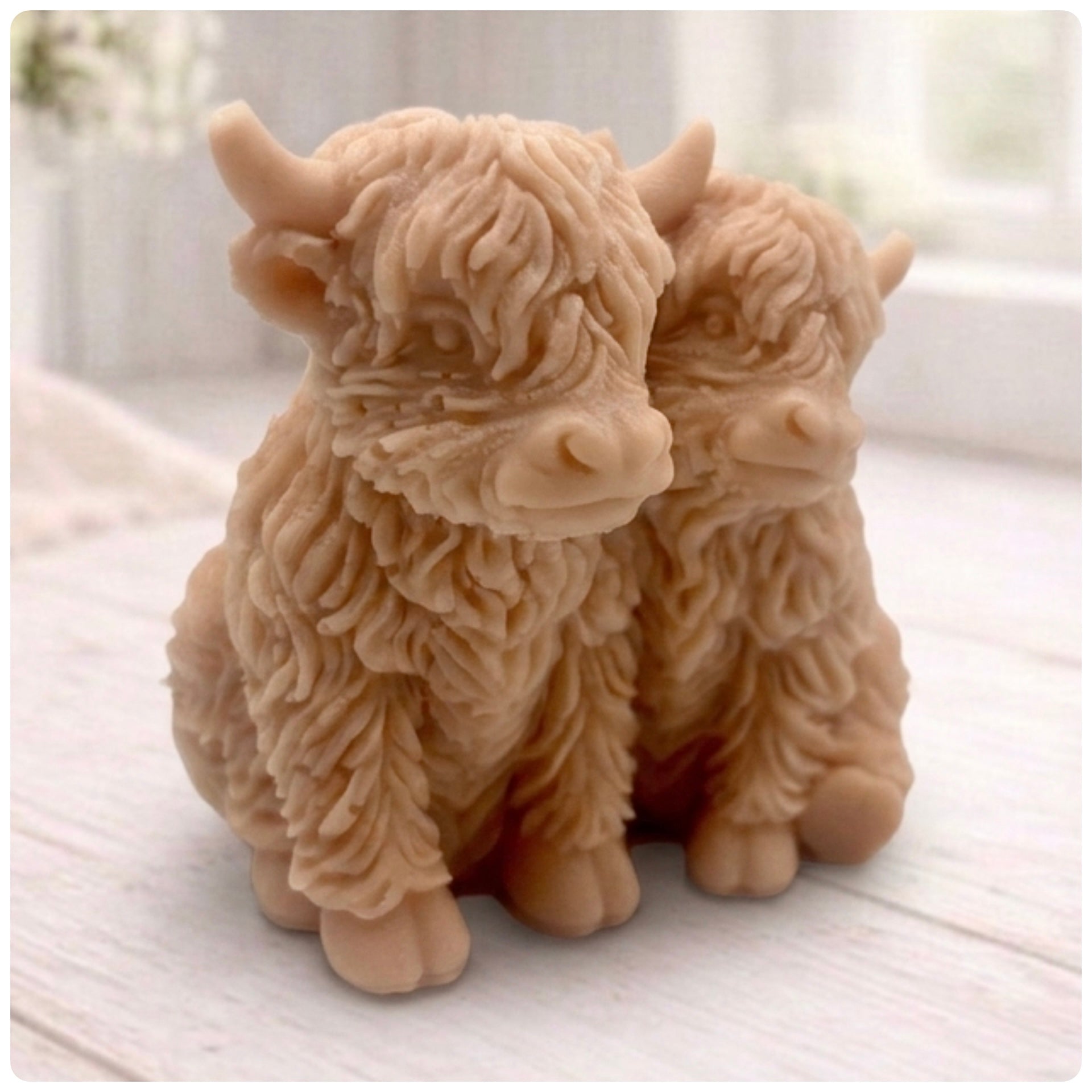 Handmade soap featuring two Highland cows with textured, shaggy fur sitting side by side on a light wooden surface with a softly indoor background.