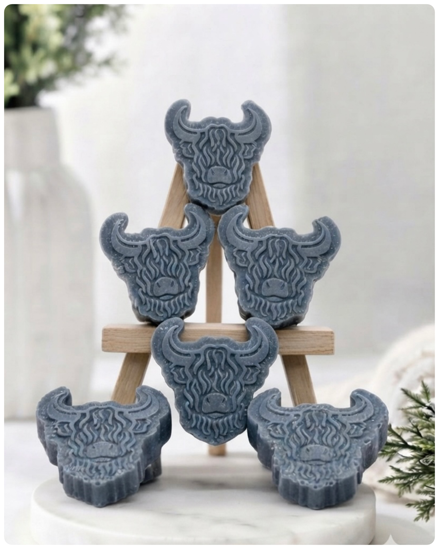 A collection of six handmade vegan soaps each intricately sculpted into the head of a shaggy Highland cow with long horns and a thick, textured coat, arranged in a pyramid on and around a small wooden easel.