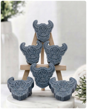 Load image into Gallery viewer, A collection of six handmade vegan soaps each intricately sculpted into the head of a shaggy Highland cow with long horns and a thick, textured coat, arranged in a pyramid on and around a small wooden easel.
