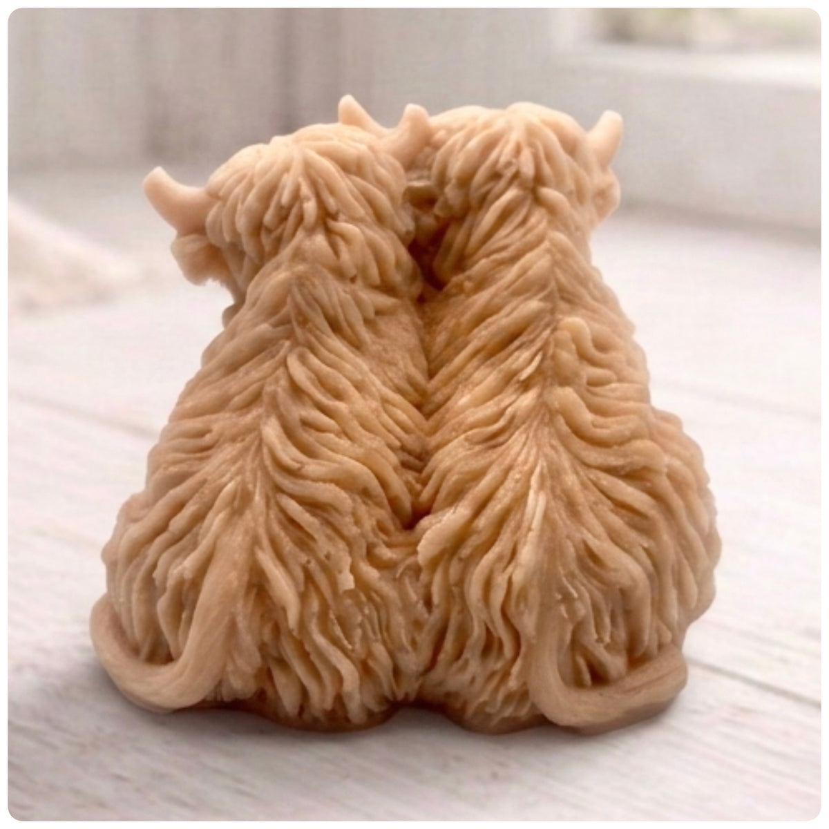 Handmade soap featuring two Highland cows with textured, shaggy fur sitting side by side on a light wooden surface with a softly indoor background.