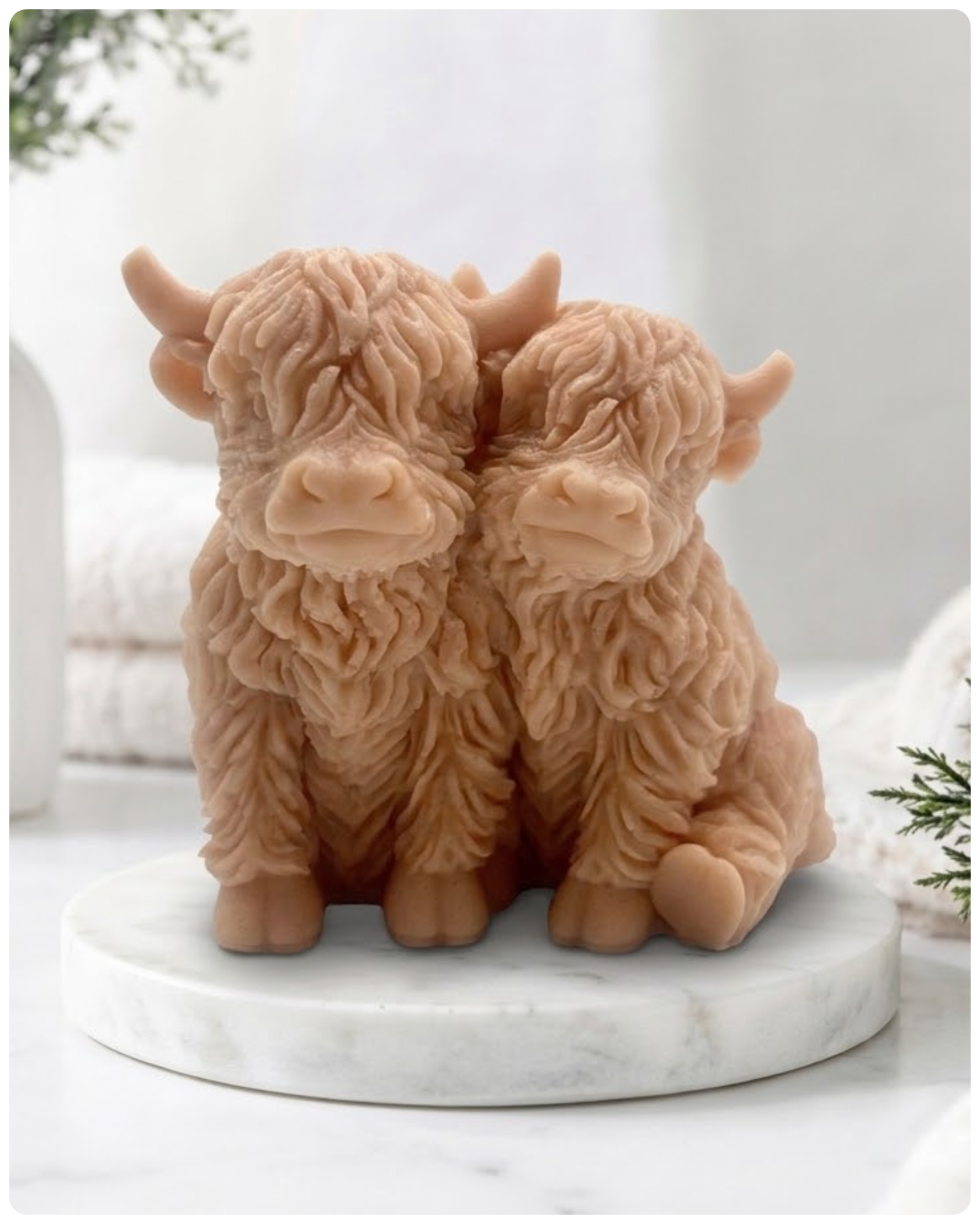 Handmade soap featuring two Highland cows with textured, shaggy fur sitting side by side on a light wooden surface with a softly indoor background.