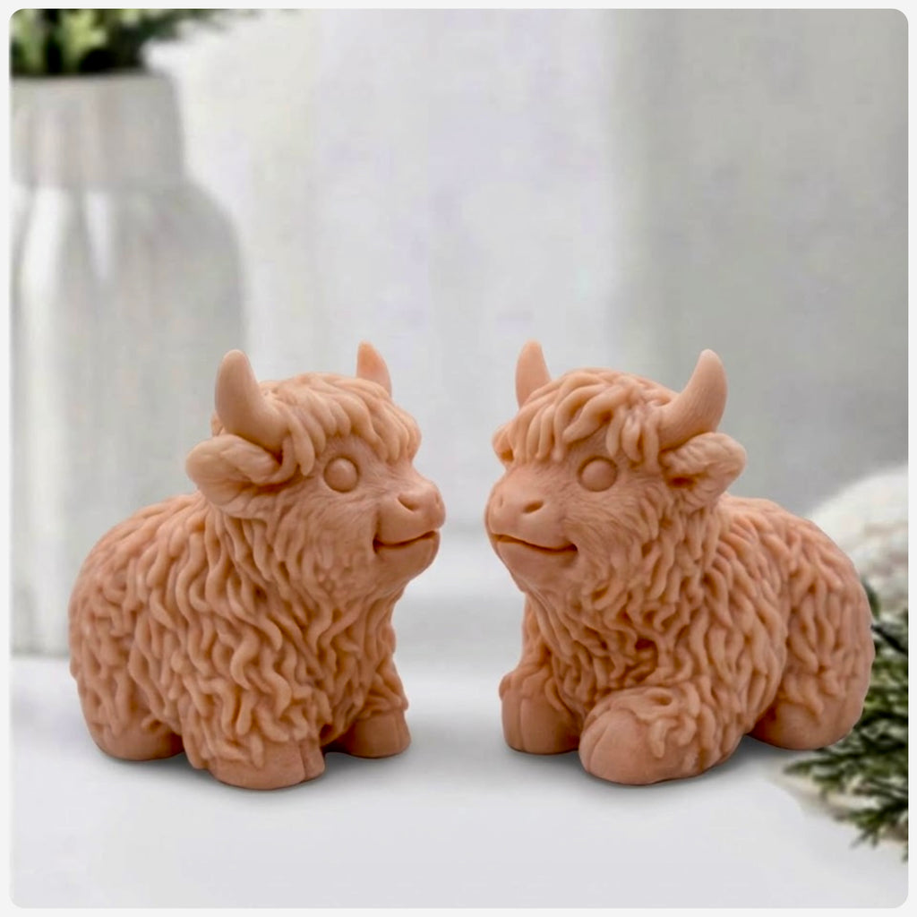 Two Red French Clay coloured Highland cow soaps are sitting side-by-side and facing each other, on a surface with a blurry background of a vase and a towel.