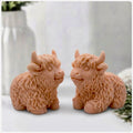 Two Red French Clay coloured Highland cow soaps are sitting side-by-side and facing each other, on a surface with a blurry background of a vase and a towel.