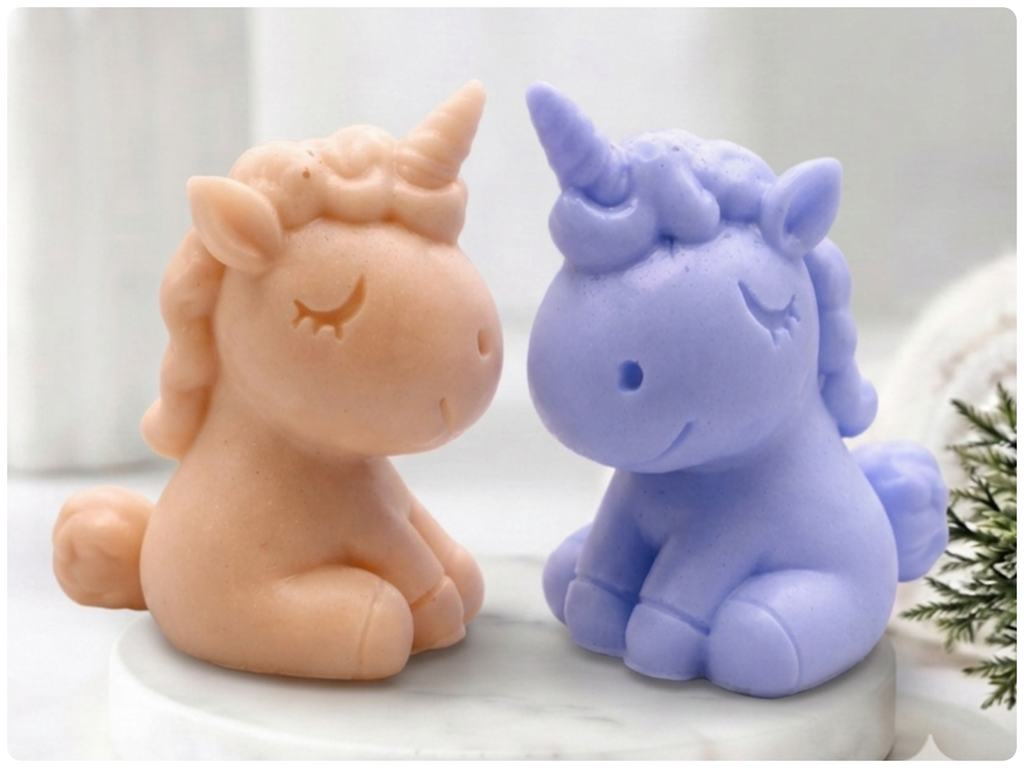 Two unicorn soaps sitting side by side on a white marble-like surface. One unicorn is peachy beige and the other is light lavender purple.